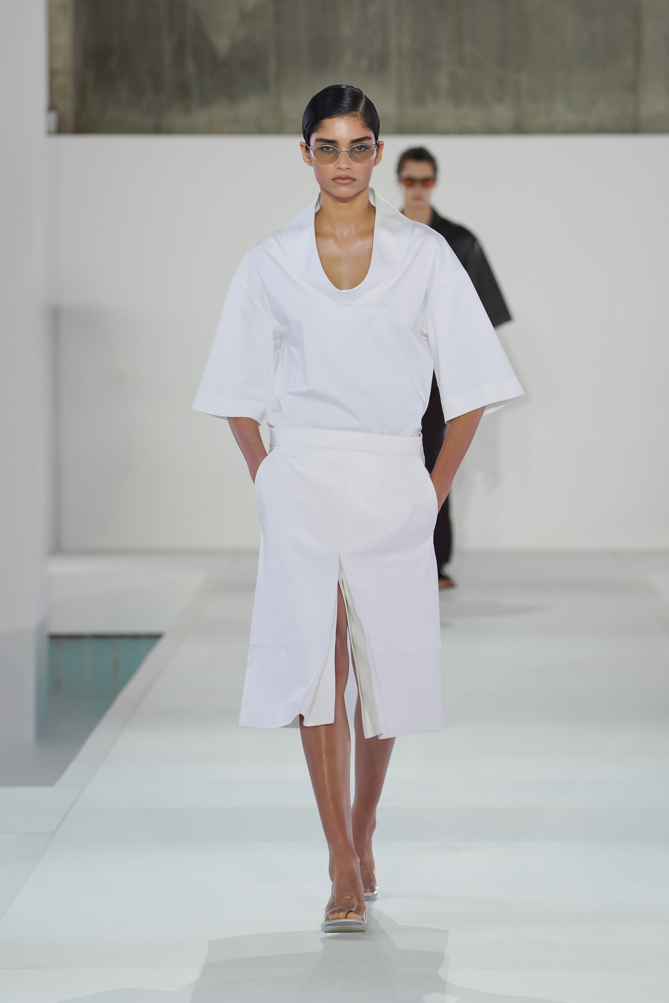 Model in white short-sleeved outfit and sunglasses walks runway with another model in the background, set against a minimalist backdrop.