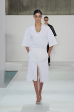 Placeholder for: Model in white short-sleeved outfit and sunglasses walks runway with another model in the background, set against a minimalist backdrop.