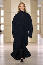 Placeholder for: Model wearing a long, dark navy overcoat with a matching turtleneck, walking on a minimalist runway with neutral-colored panels.