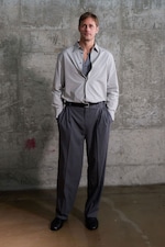 Placeholder for: Man in a partially unbuttoned gray shirt and dark trousers stands against a textured concrete wall, with hands in pockets.