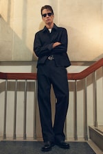 Placeholder for: Man in black outfit and sunglasses stands confidently on stairs with arms crossed. Light from above casts shadows.