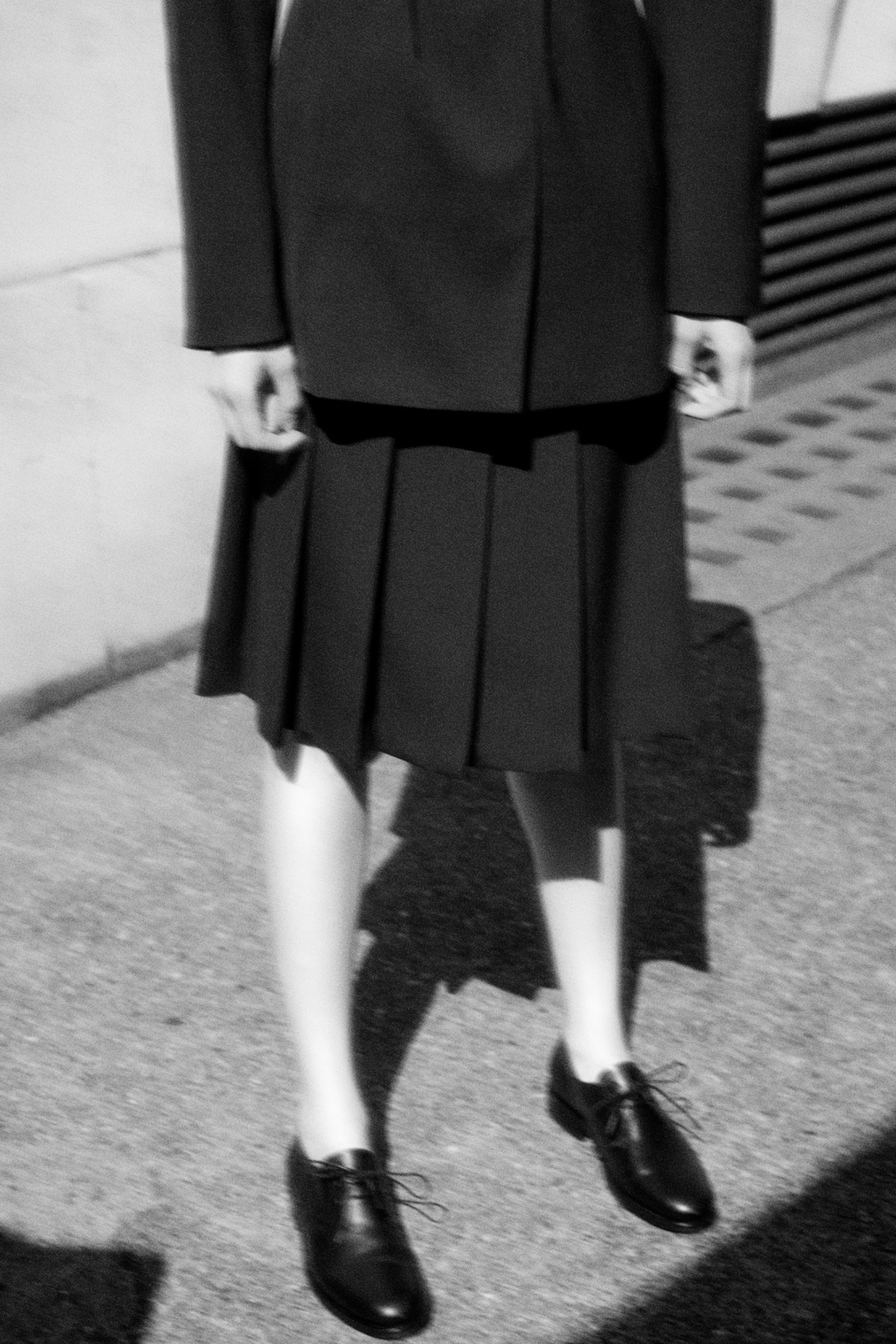 Black and white image of a person in a dark pleated skirt, blazer, and black shoes, standing on a textured pavement.