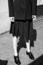 Placeholder for: Black and white image of a person in a dark pleated skirt, blazer, and black shoes, standing on a textured pavement.