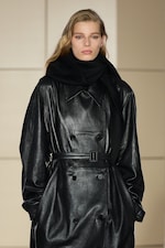 Placeholder for: Model wearing a black leather trench coat with a wide scarf, hands in pockets, light background.