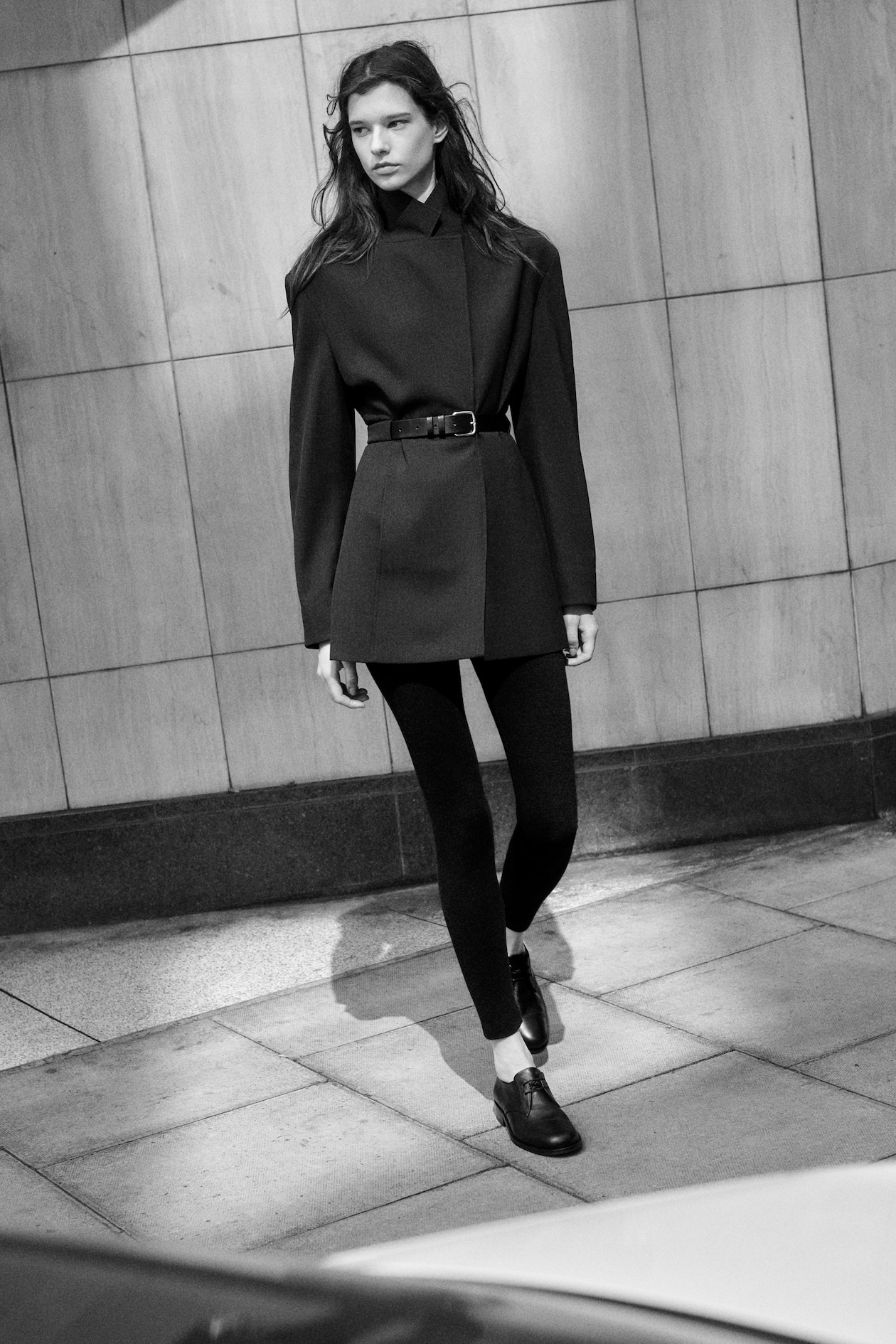 Black and white image of a person in a belted jacket and leggings, walking on a tiled sidewalk against a stone wall.