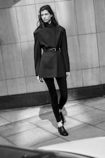 Placeholder for: Black and white image of a person in a belted jacket and leggings, walking on a tiled sidewalk against a stone wall.
