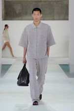 Placeholder for: A model on a runway wearing a light gray short-sleeve suit with black sandals, carrying a black bag. Another model is in the background.