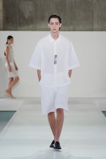 Placeholder for: Model in oversized white shirt and shorts walks runway; another model in the background wears a sheer white outfit.