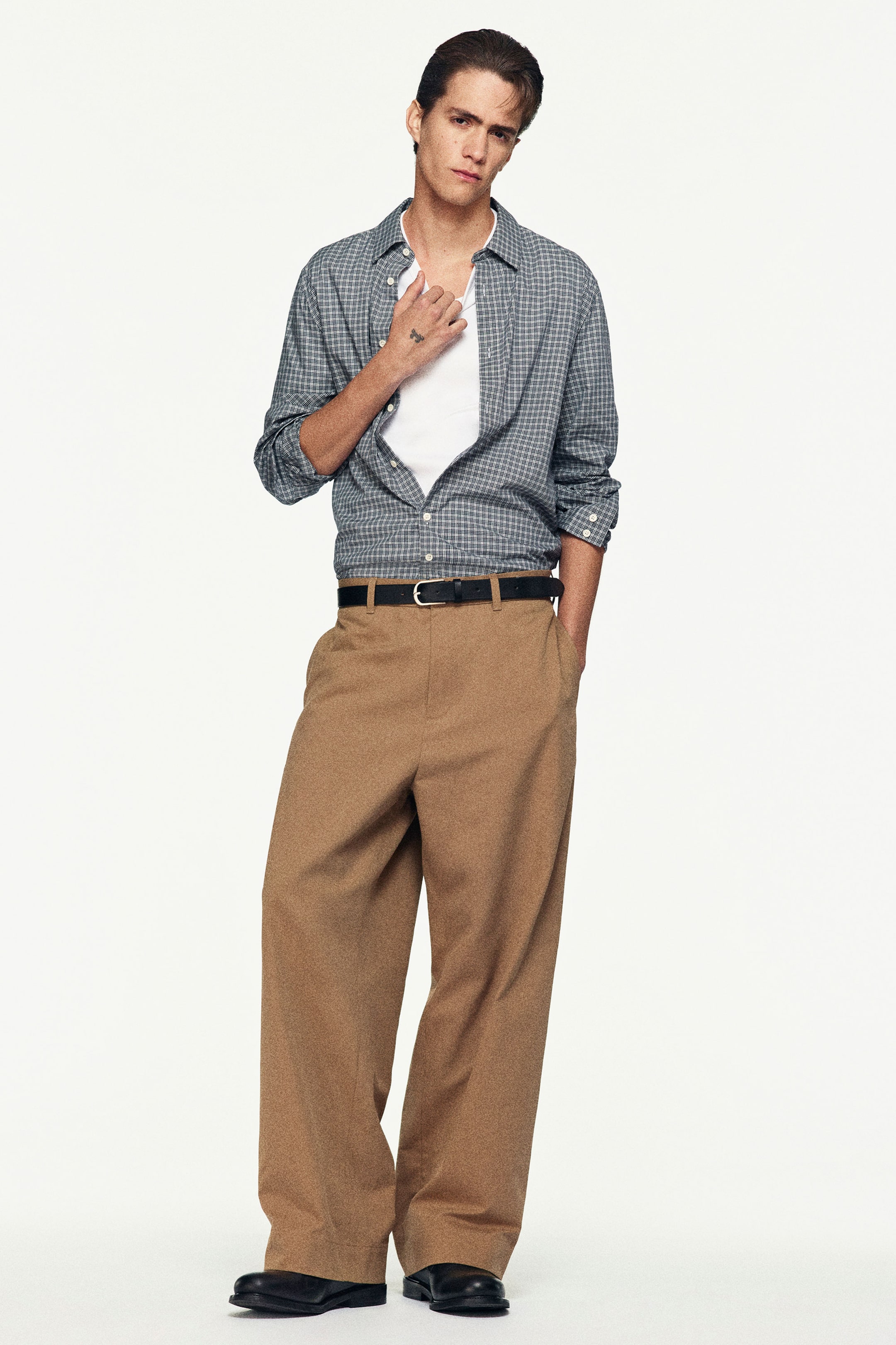 Person wearing a checkered shirt over a white top and brown wide-leg pants, standing with one hand in pocket against a plain background.