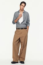 Placeholder for: Person wearing a checkered shirt over a white top and brown wide-leg pants, standing with one hand in pocket against a plain background.