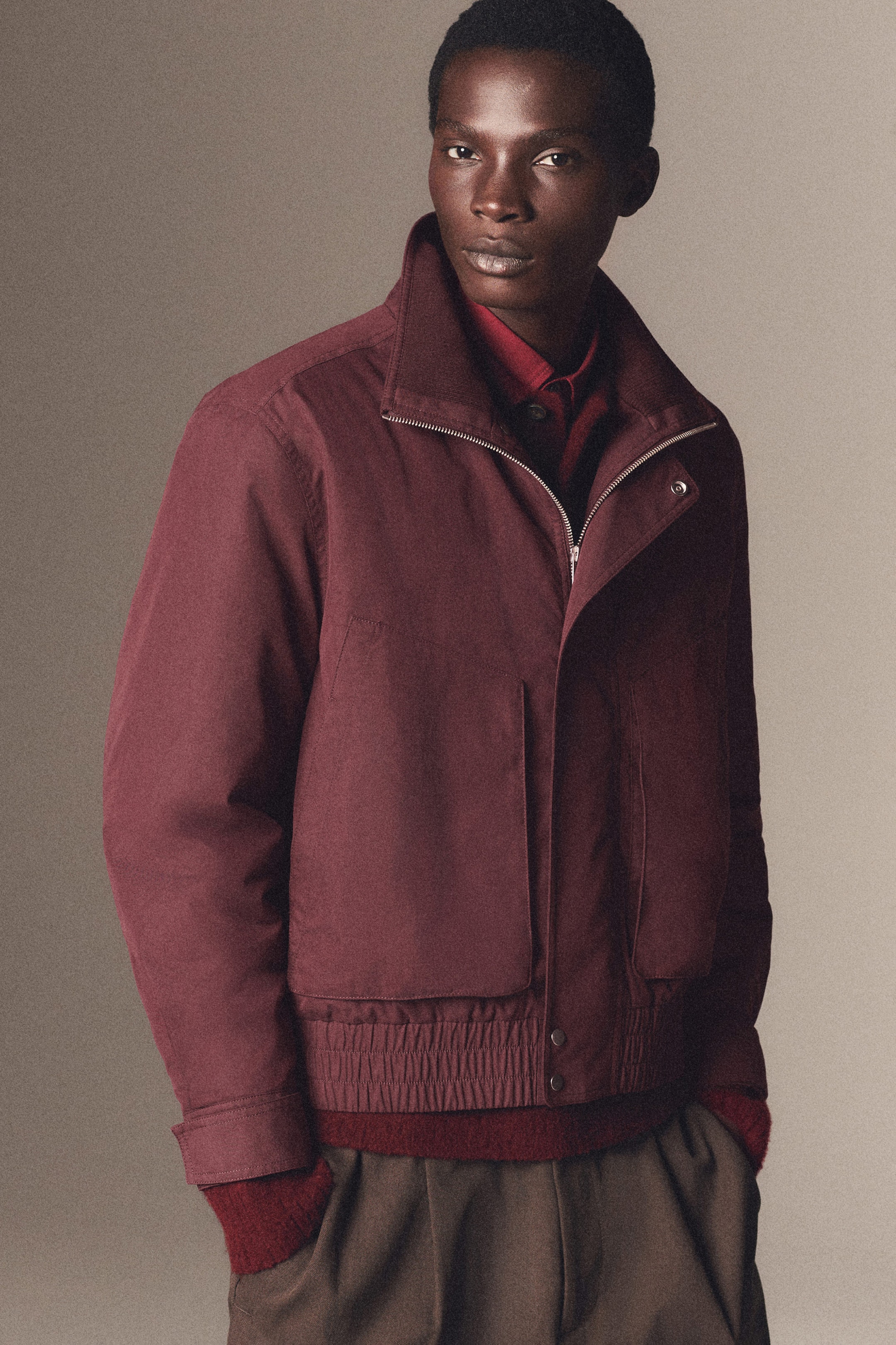 Person wearing a burgundy jacket over a red sweater and brown pants, standing against a neutral background.
