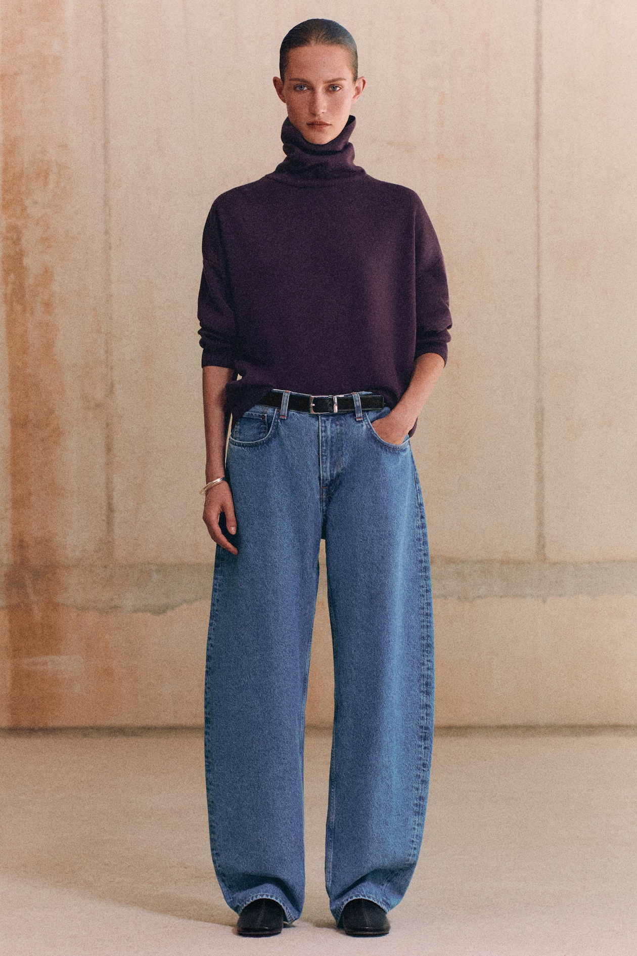 A person wearing a dark turtleneck sweater and wide-leg blue jeans, standing against a neutral background.