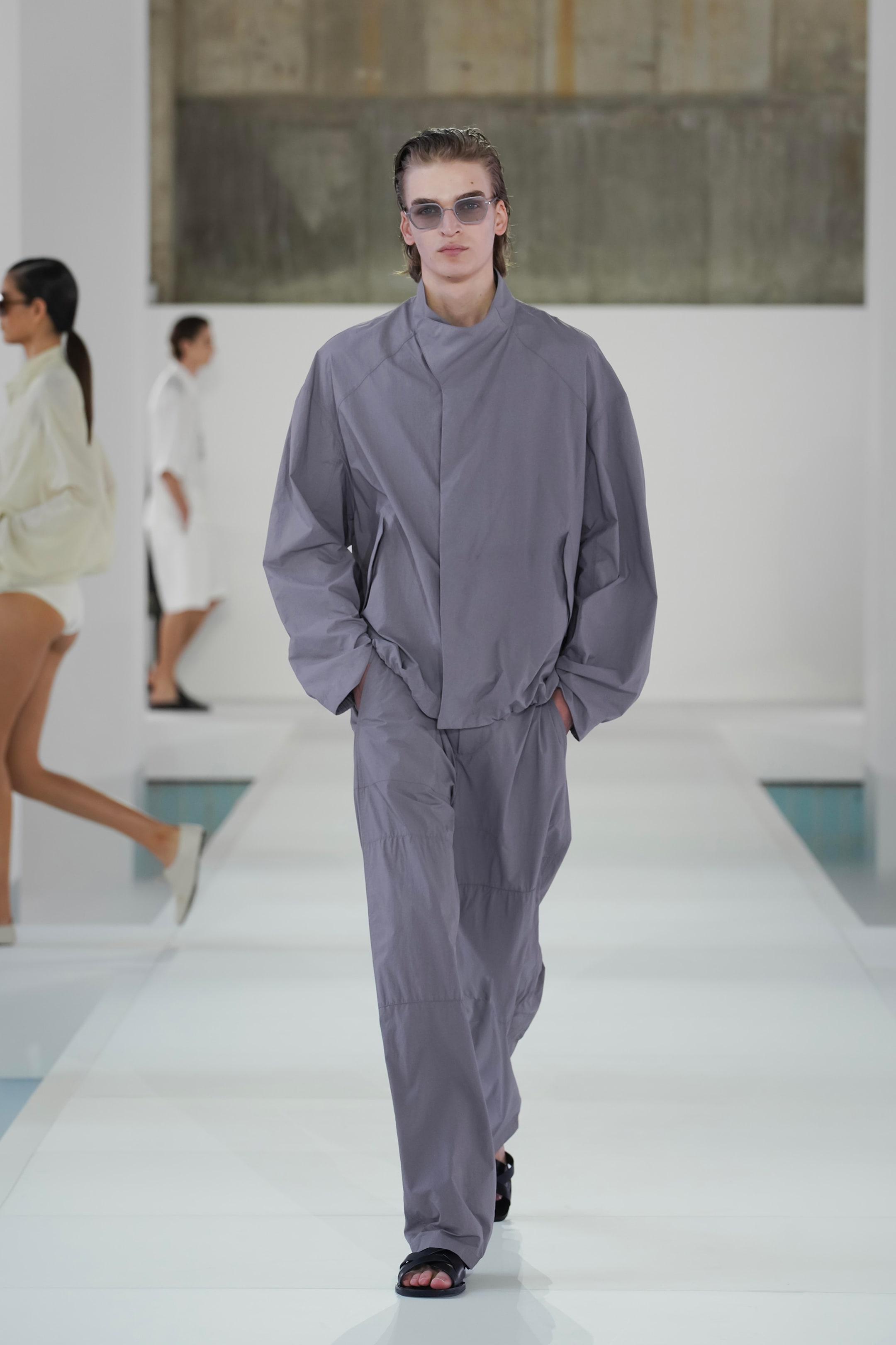 Model wearing a loose-fitting gray outfit and sunglasses walks on a runway, with other models in similar minimalist clothing behind.