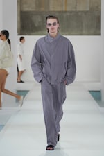 Placeholder for: Model wearing a loose-fitting gray outfit and sunglasses walks on a runway, with other models in similar minimalist clothing behind.