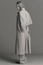 Placeholder for: Person in a long, belted coat and trousers, facing sideways against a neutral background in a monochrome setting.