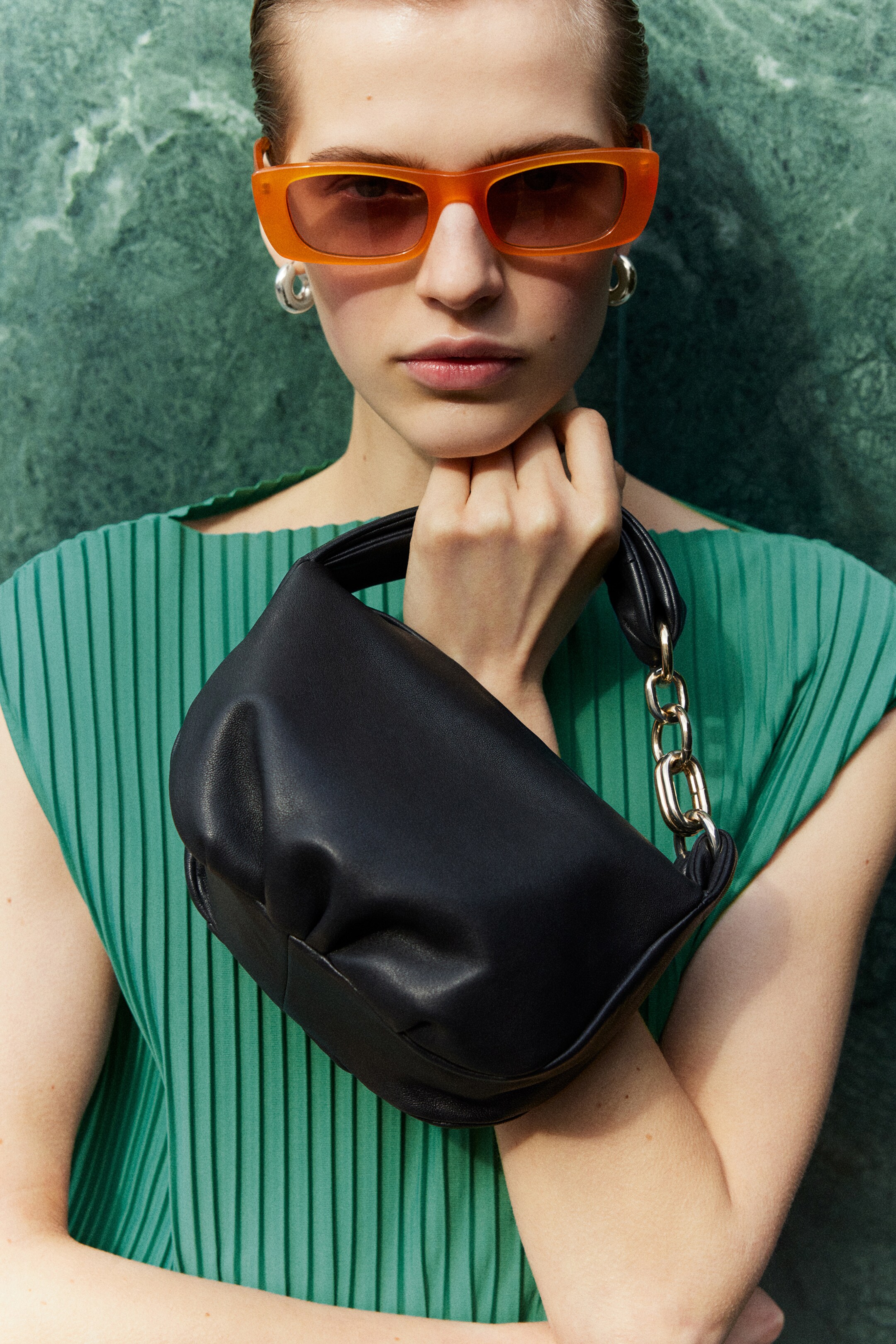 Person wearing orange sunglasses, green pleated top holding black leather bag