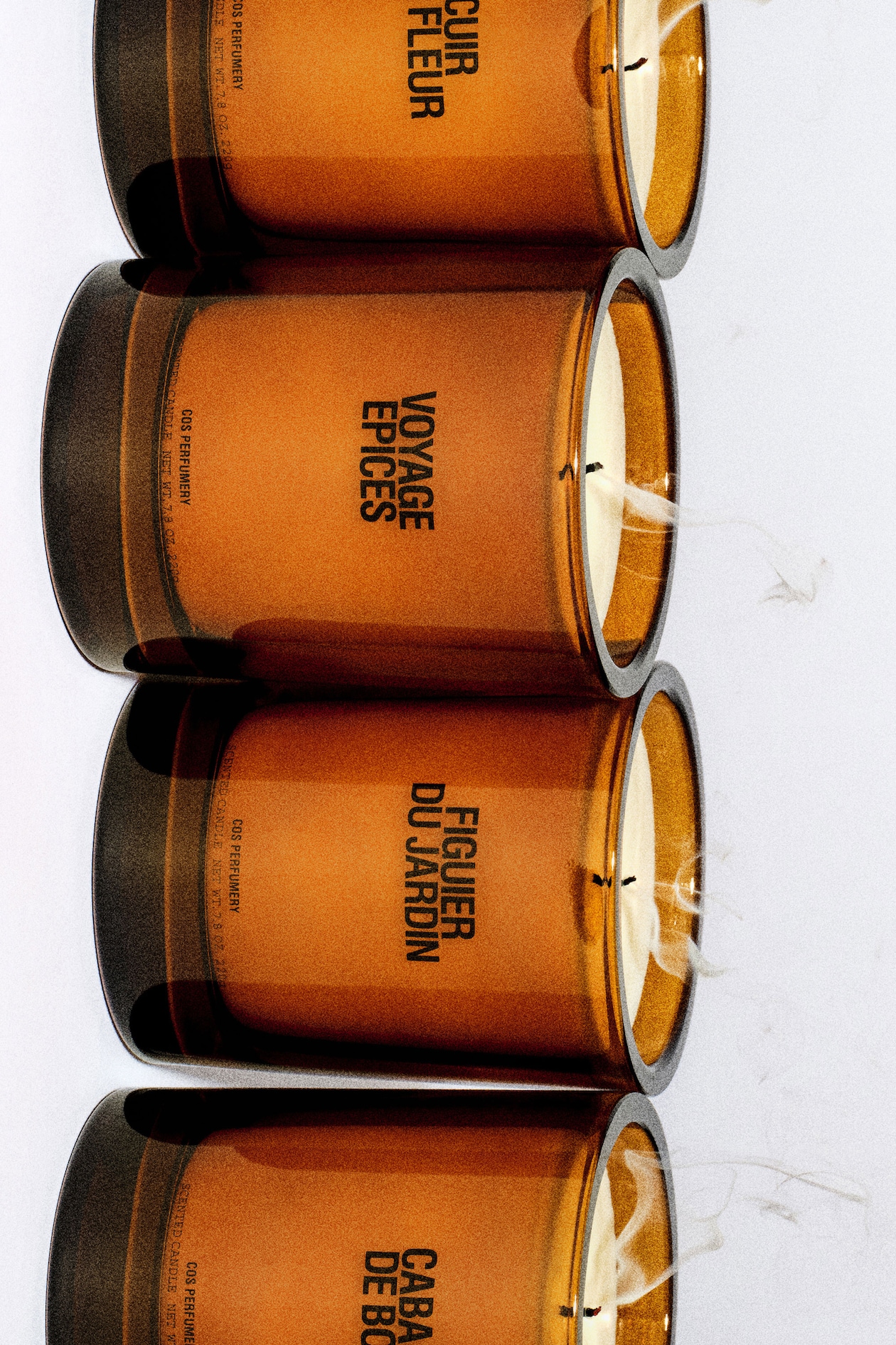 Four amber-colored candle jars with white wicks on a white surface, labeled with different fragrance names.