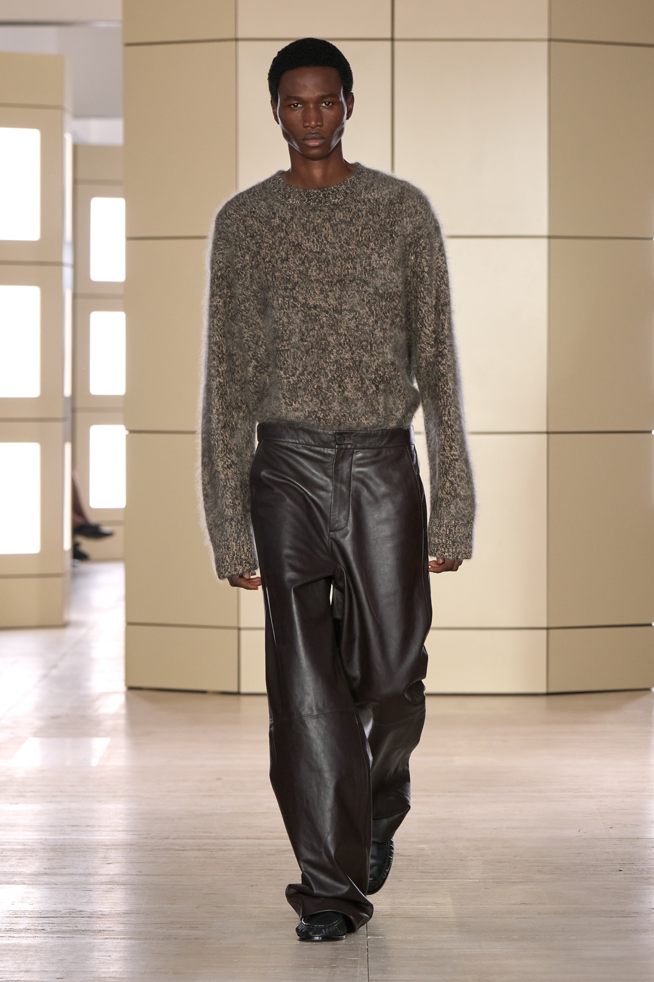 Model walking in a fashion show wearing a fuzzy gray sweater and dark brown leather pants. The setting has a modern interior design.