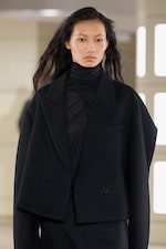 Placeholder for: A model wearing an oversized black blazer and sheer turtleneck walks on a runway.