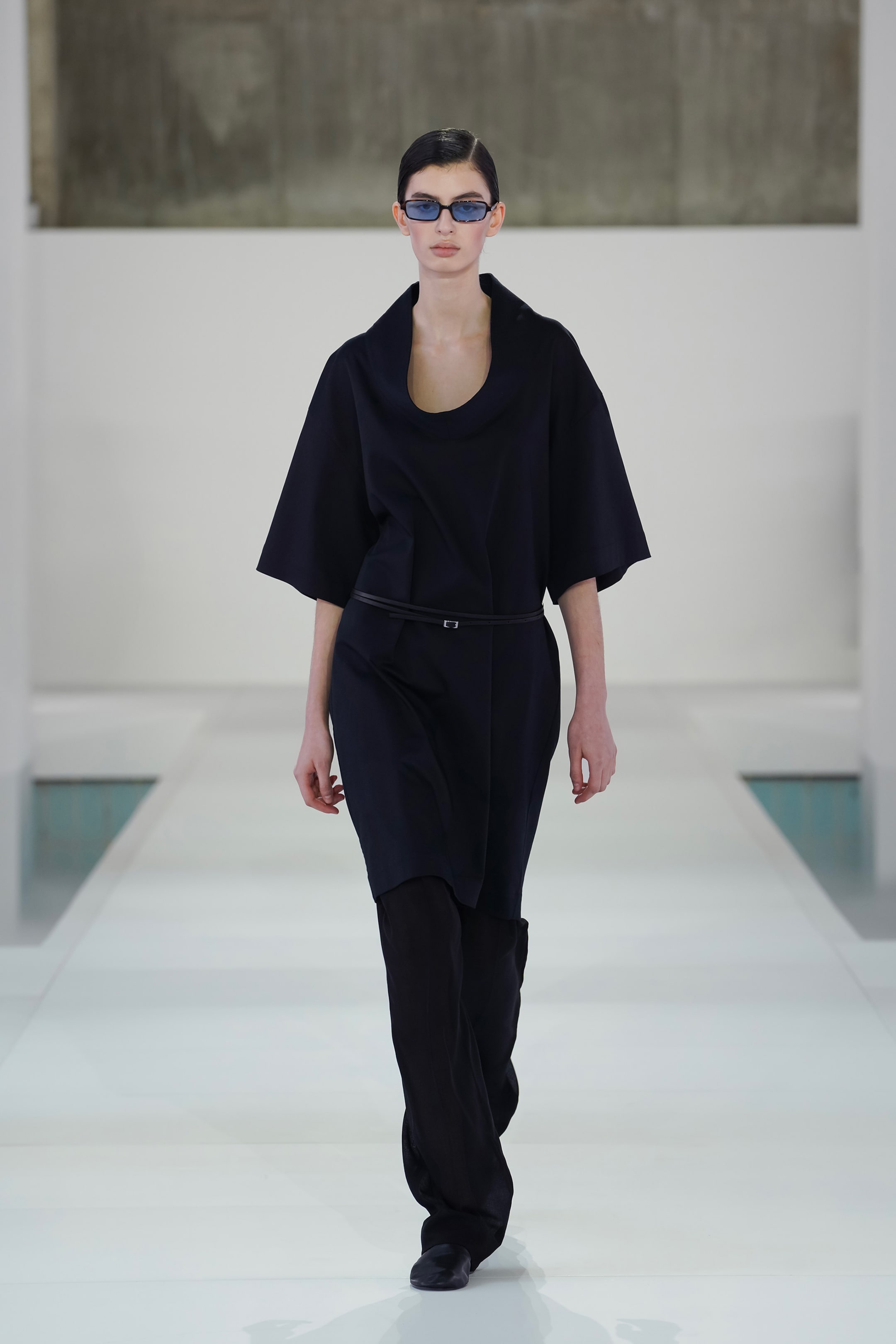 Model in a sleek black outfit and sunglasses walking on a white runway with a minimalist background.