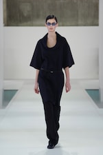 Placeholder for: Model in a sleek black outfit and sunglasses walking on a white runway with a minimalist background.