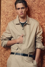 Placeholder for: Young man in a beige jacket leans against a cork wall, with one hand in his pocket. He has short hair and a neutral expression.
