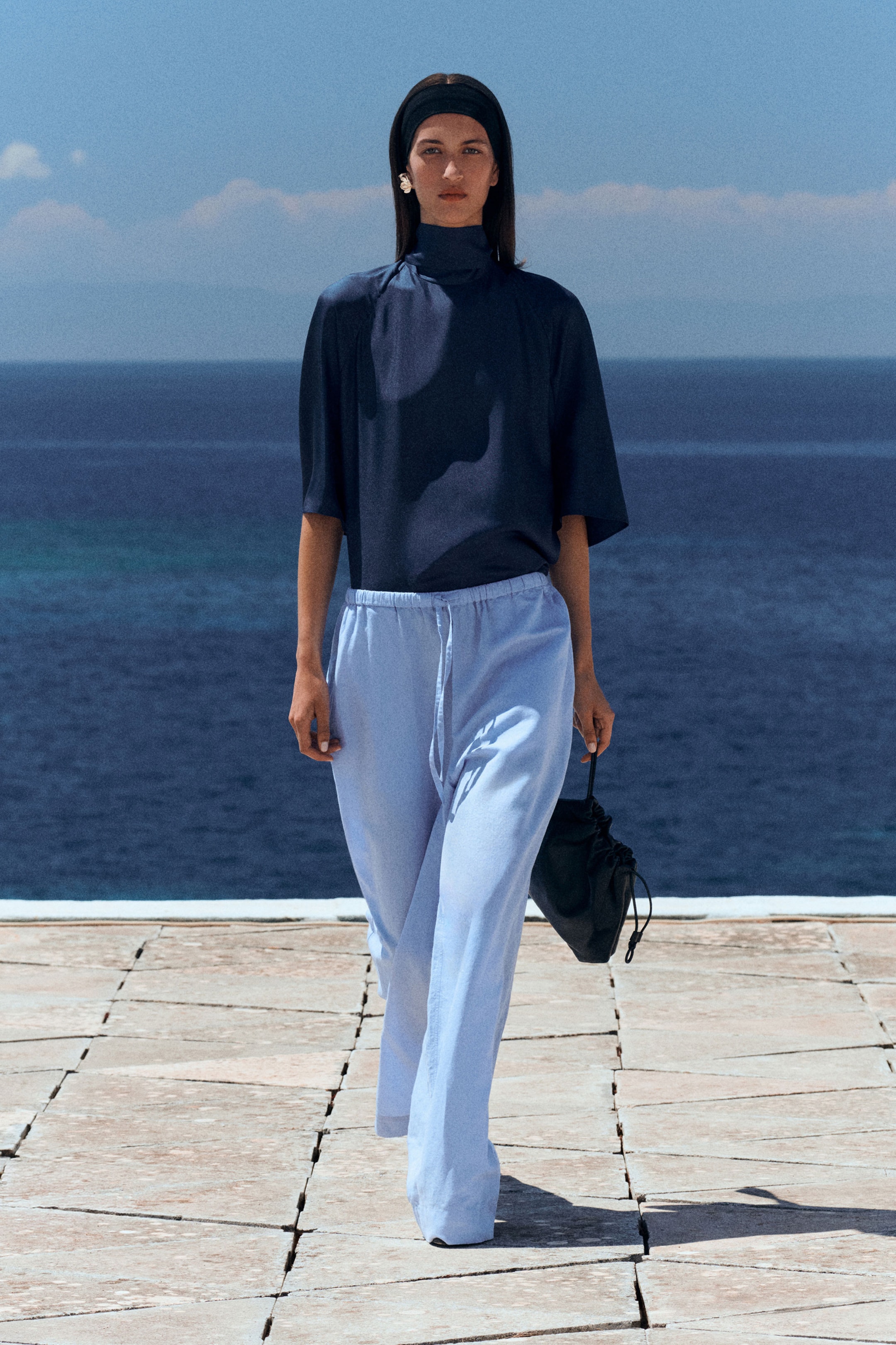 Woman in navy top and light blue pants walks on a tiled path by the sea, holding a small black bag, against a clear blue sky.