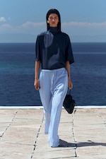 Placeholder for: Woman in navy top and light blue pants walks on a tiled path by the sea, holding a small black bag, against a clear blue sky.