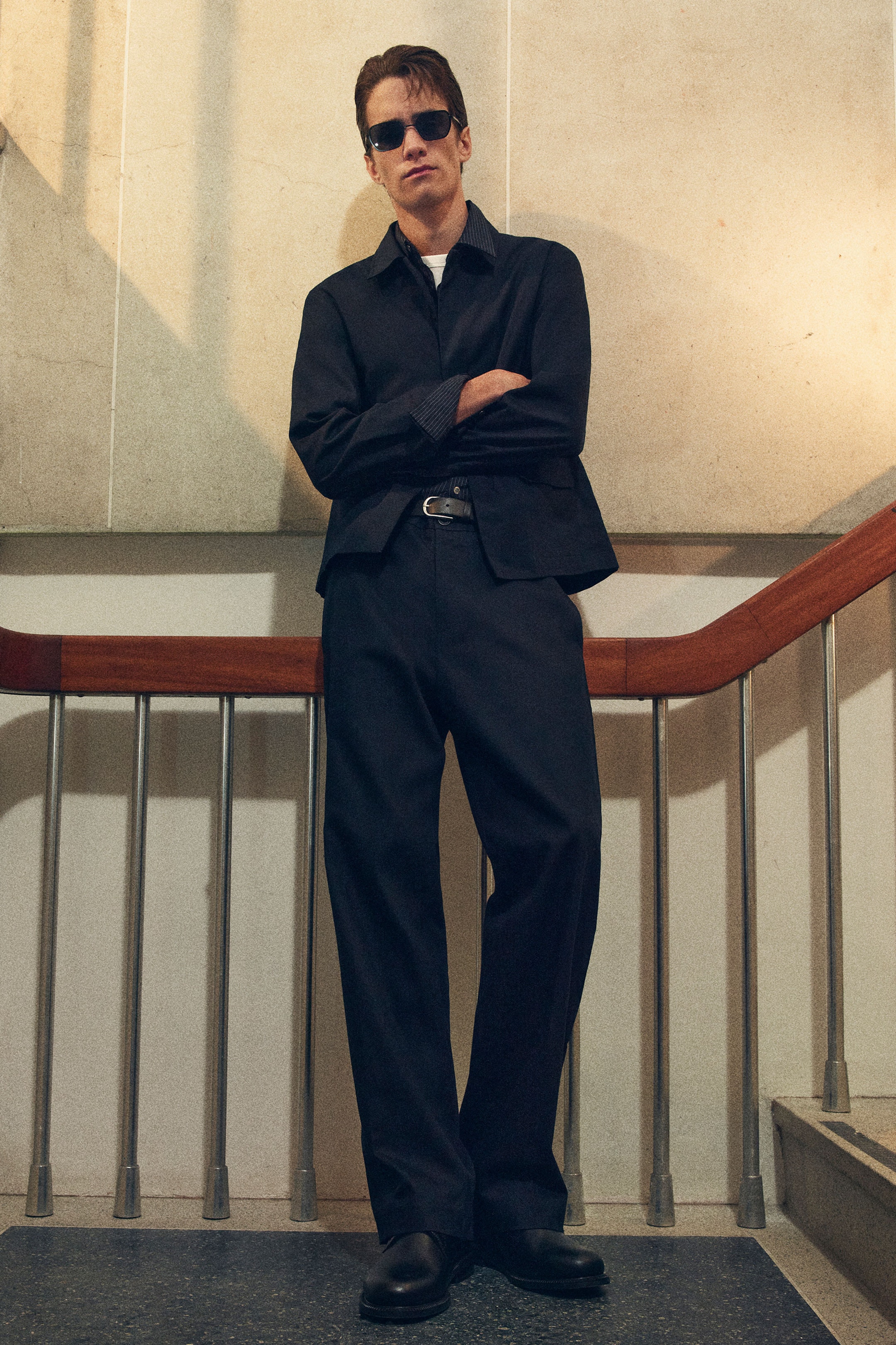 Man in dark outfit and sunglasses stands confidently on stairs, arms crossed. Steps and metal railing visible in the background.
