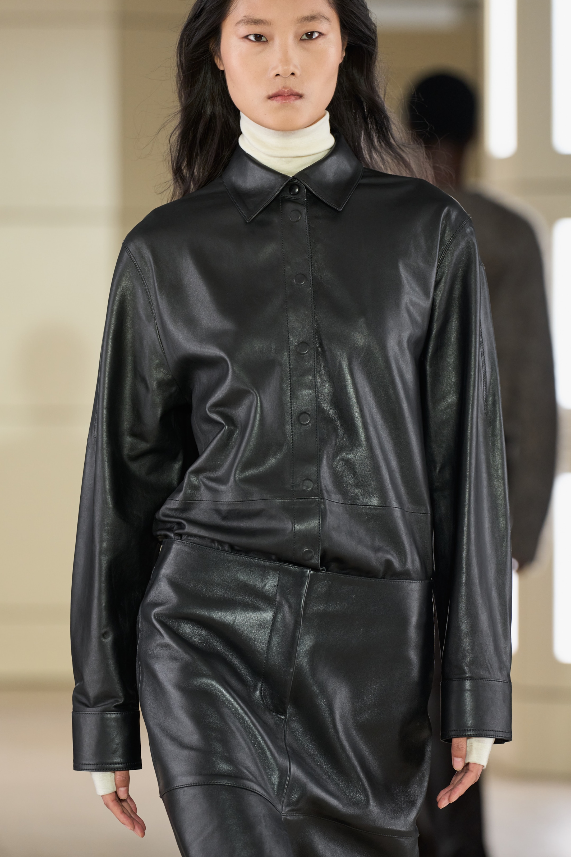 Model wearing a black leather outfit with a white turtleneck underneath, walking on a runway.