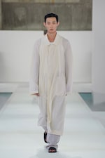 Placeholder for: Model in a long, white, sheer coat walks on a white runway. They wear gray pants and black sandals, with a neutral expression.