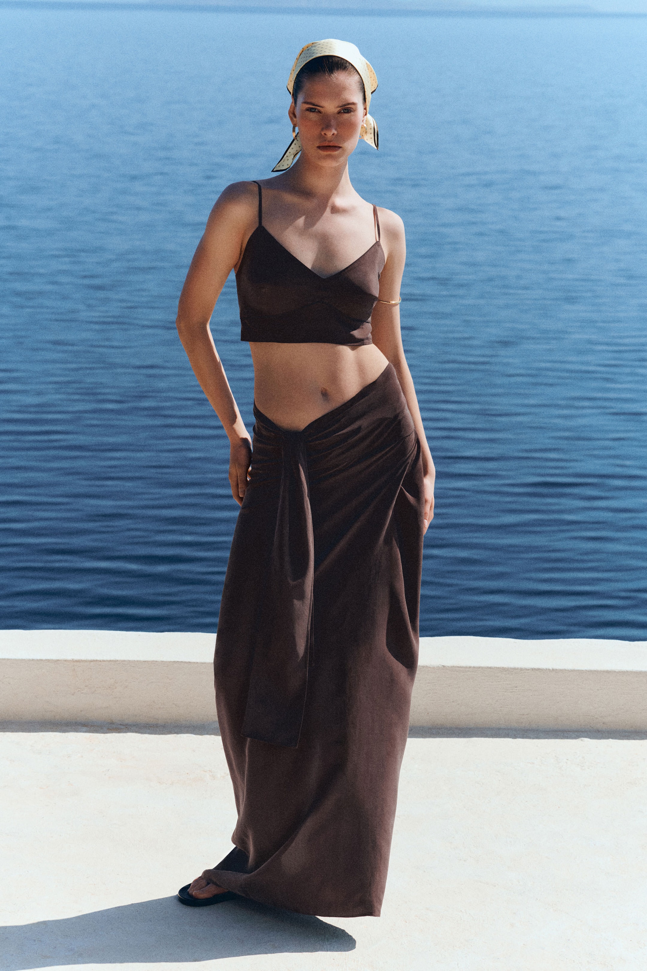 Woman in a brown crop top and skirt stands by the sea, wearing a headscarf, against a backdrop of deep blue water.