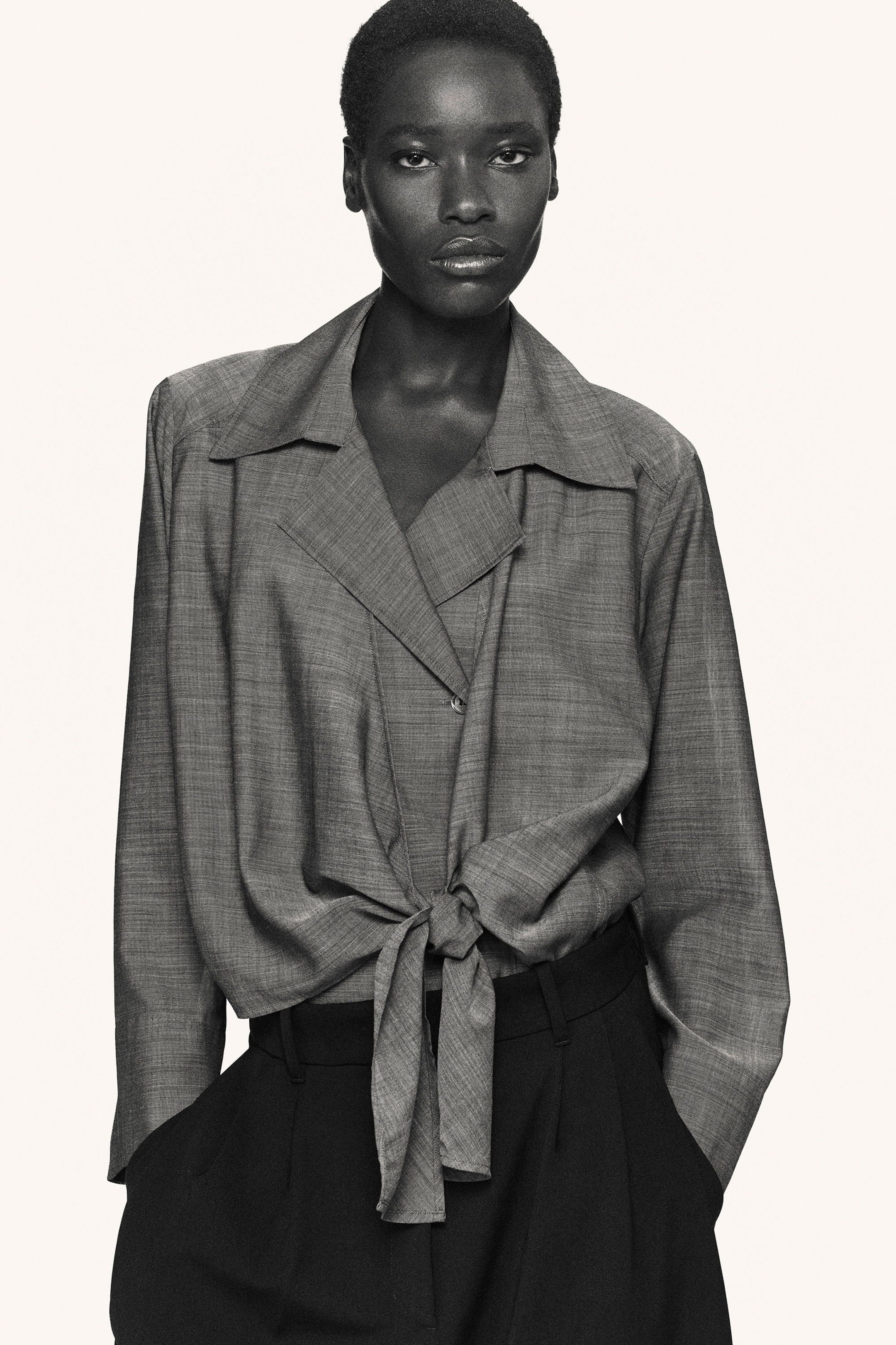 Person wearing a light gray long-sleeve blouse tied at the waist and dark trousers, standing against a plain background.