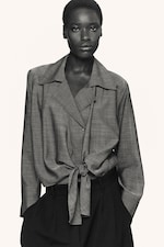 Placeholder for: Person wearing a light gray long-sleeve blouse tied at the waist and dark trousers, standing against a plain background.