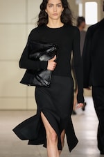 Placeholder for: A model in a black outfit with a thigh-high slit walks down a runway, holding a large black leather clutch.