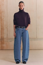 Placeholder for: Person wearing a dark turtleneck sweater and wide-leg jeans stands against a beige wall.