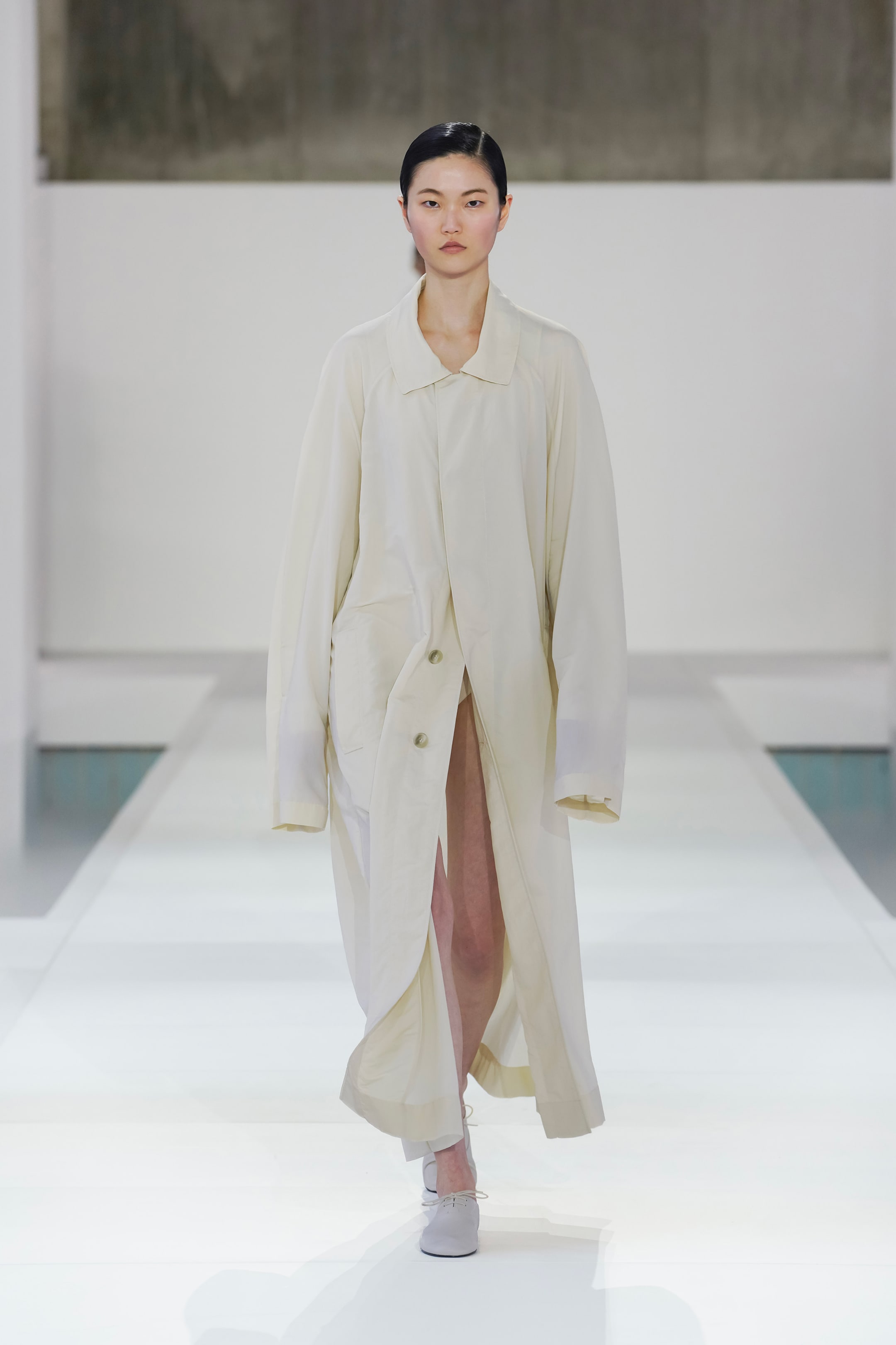 A model walks down the runway in an oversized, cream-colored coat with large buttons, set against a minimalist, light background.
