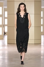 Placeholder for: Model in a black sleeveless dress with floral cutouts and pockets, walking on a wooden runway with beige paneled background.