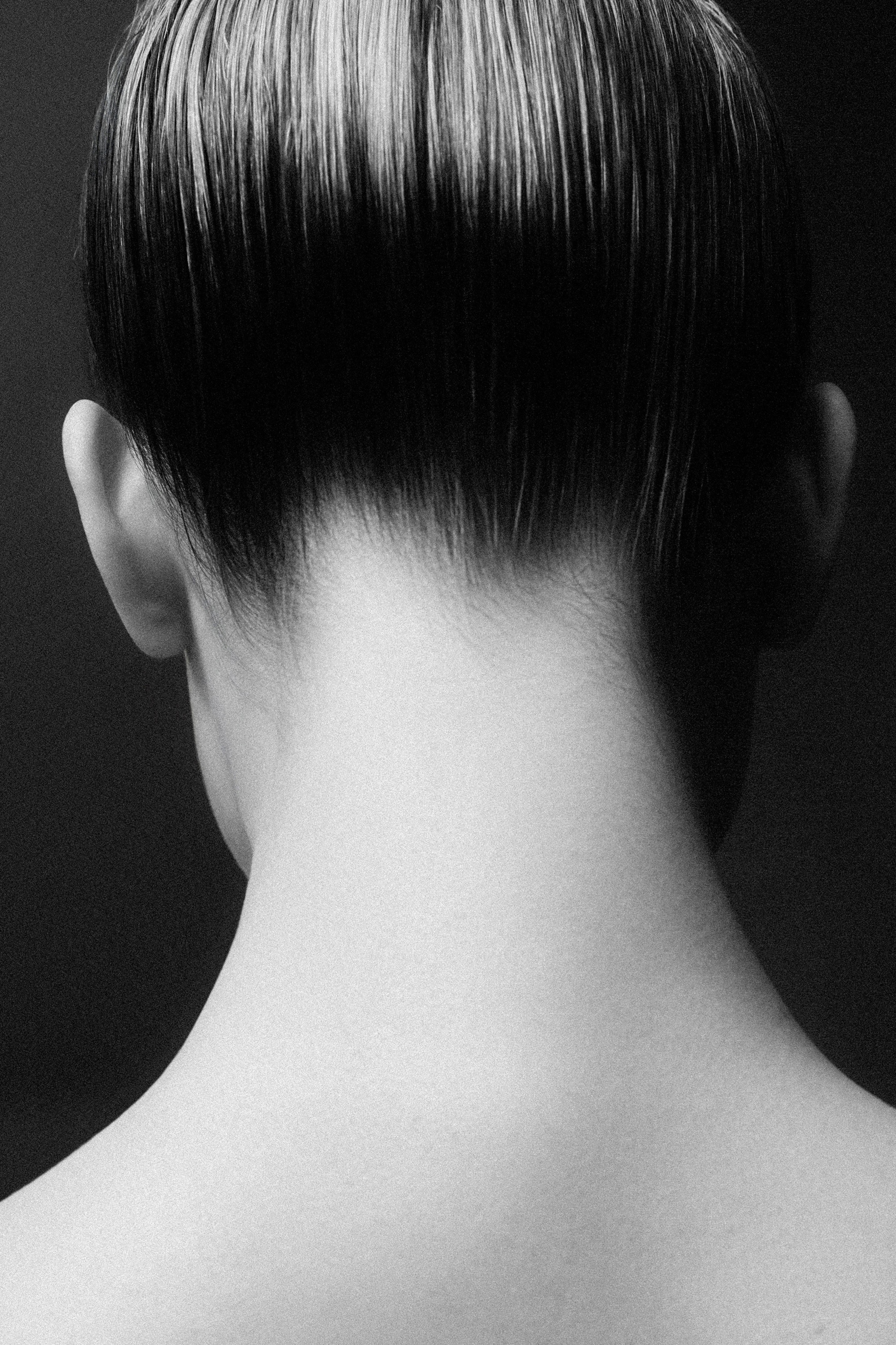 Black and white close-up of the back of a person's head and neck with neatly slicked-back hair.