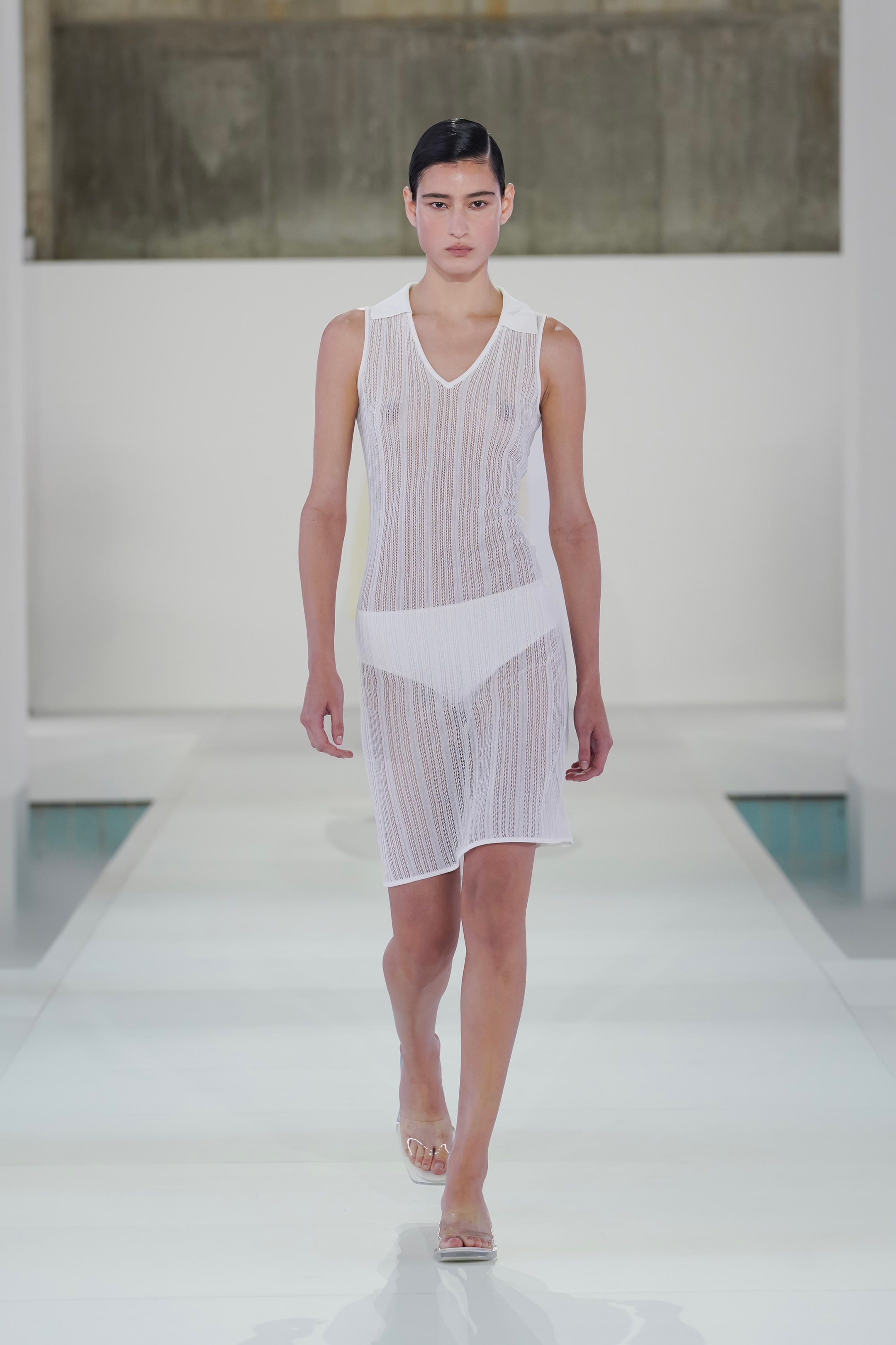Model wearing a sheer white dress with white undergarments walks confidently on a minimalist runway with a neutral backdrop.