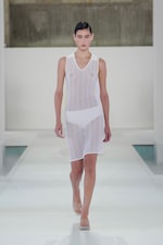 Placeholder for: Model wearing a sheer white dress with white undergarments walks confidently on a minimalist runway with a neutral backdrop.