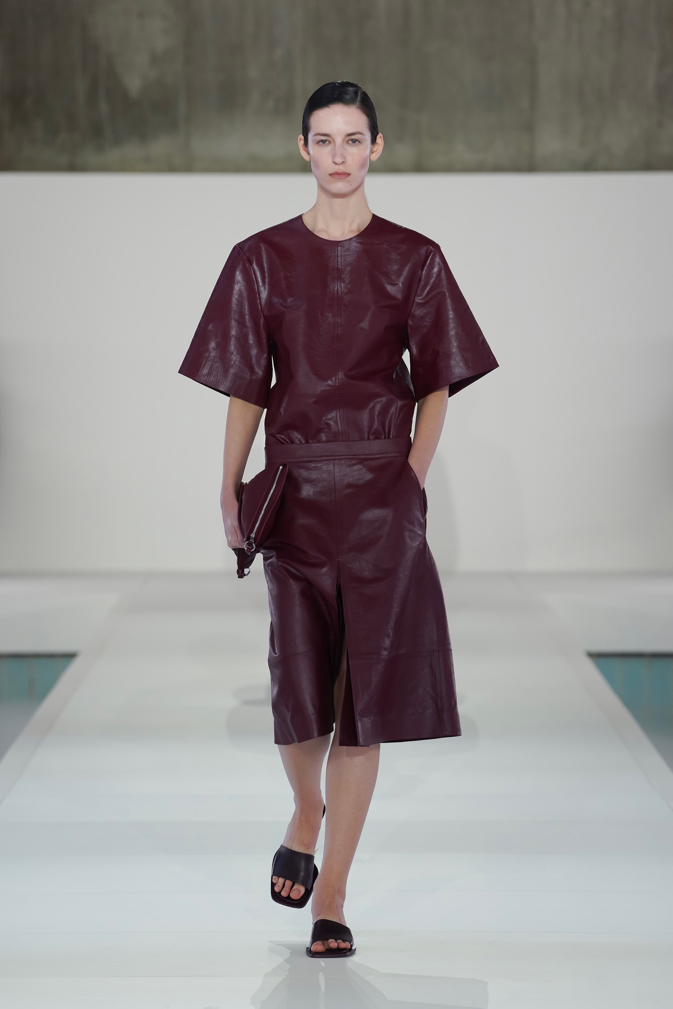 A model walks the runway wearing a dark maroon leather outfit with wide sleeves and a skirt, holding a matching clutch.