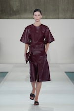Placeholder for: A model walks the runway wearing a dark maroon leather outfit with wide sleeves and a skirt, holding a matching clutch.