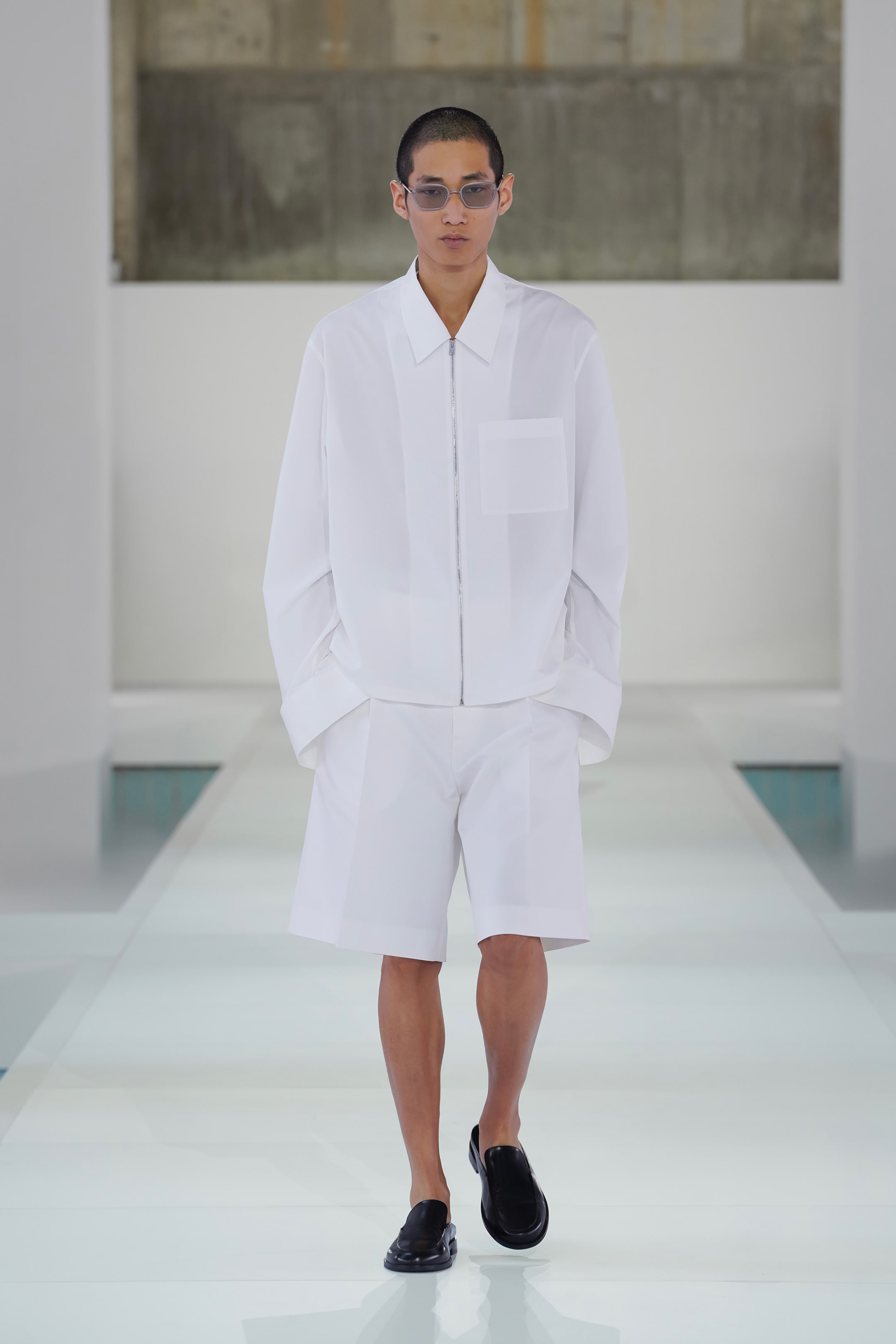 Model in white shirt and shorts walks a minimalist runway, wearing sunglasses and black shoes, against a neutral backdrop.