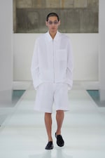Placeholder for: Model in white shirt and shorts walks a minimalist runway, wearing sunglasses and black shoes, against a neutral backdrop.