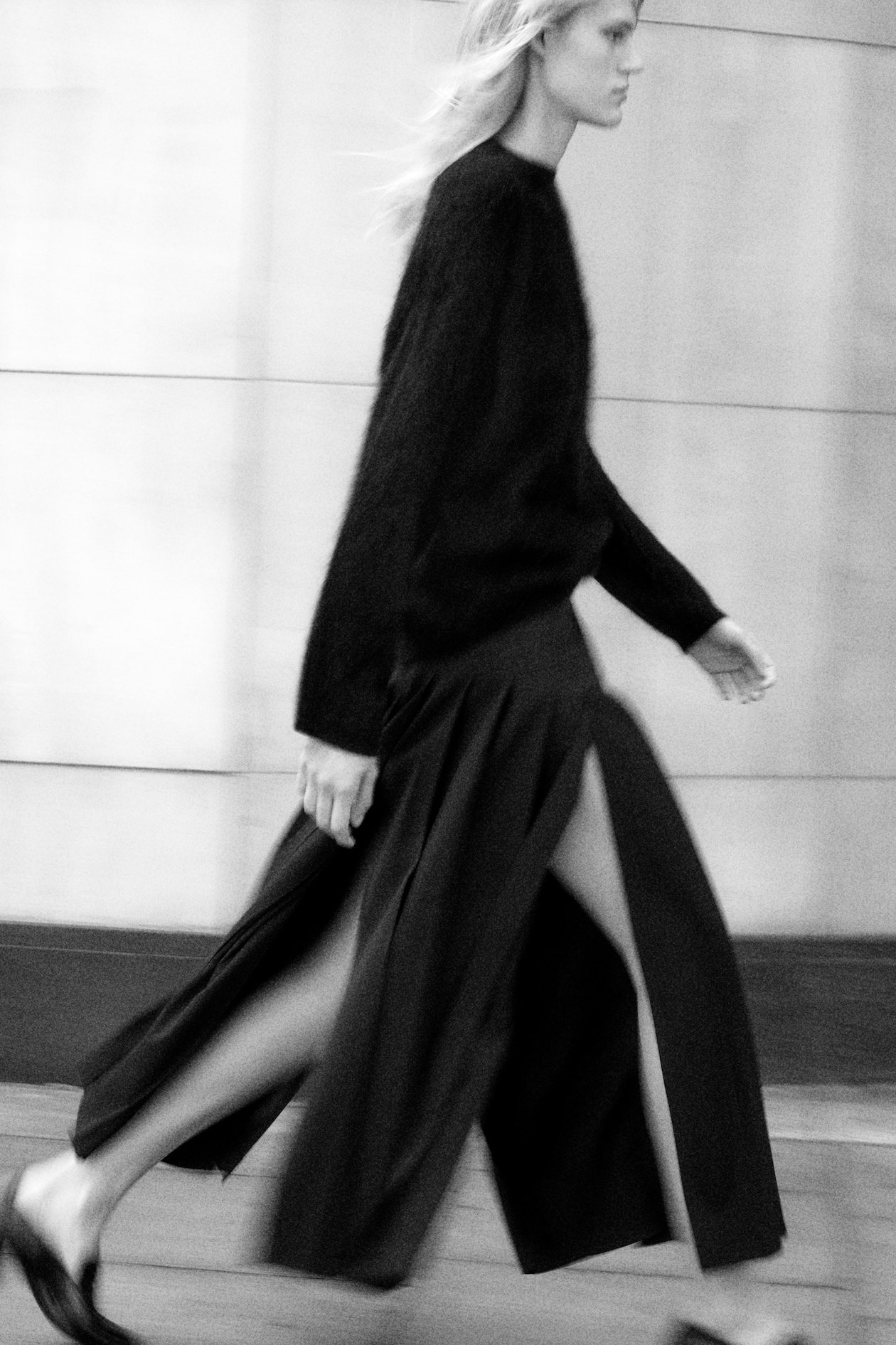 Black and white image of a person walking briskly, wearing a dark sweater and a long skirt with high slits, against a blurred background.