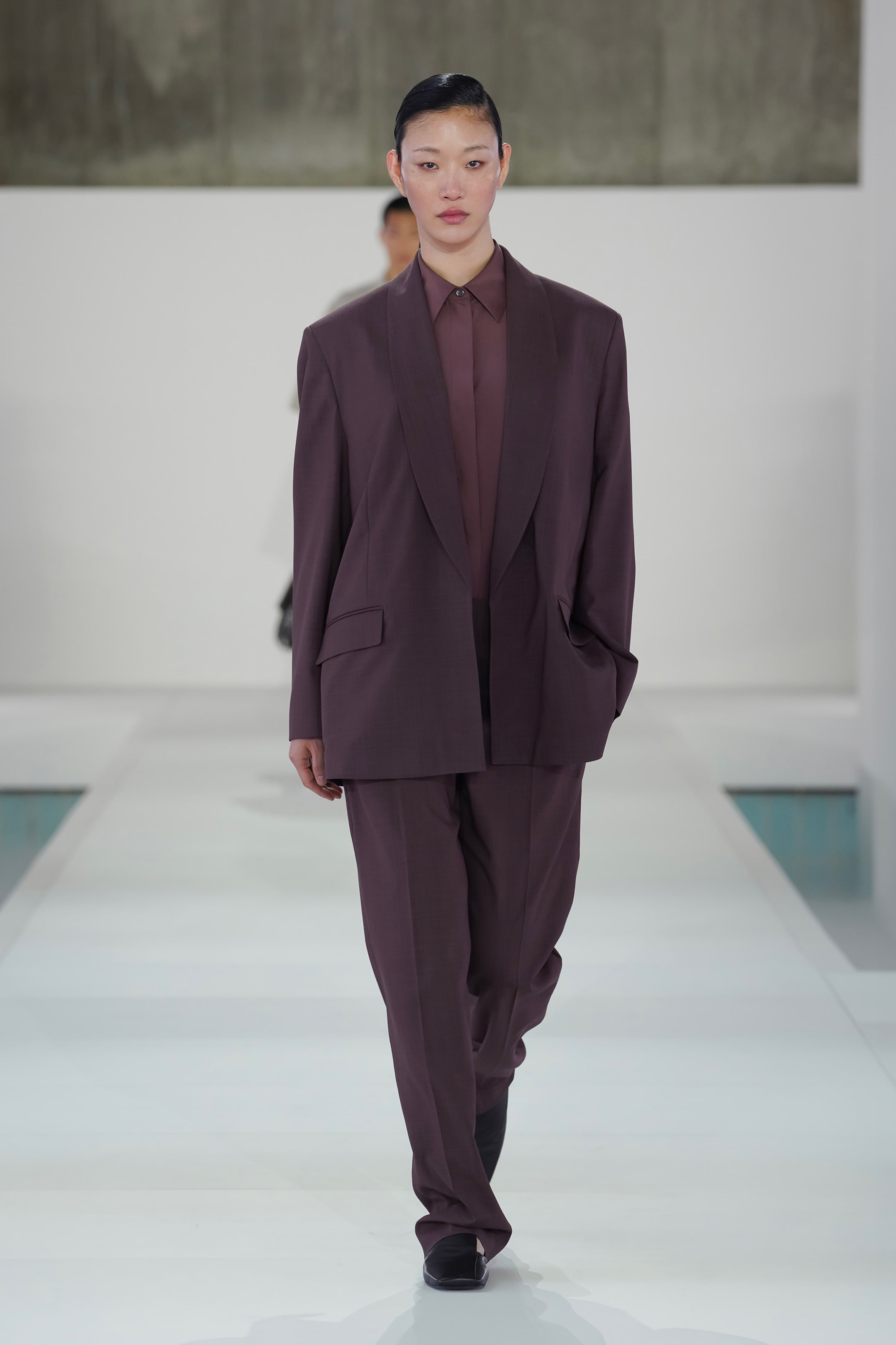 Person walking on a runway wearing an oversized brown suit with a matching shirt, against a minimalist background.