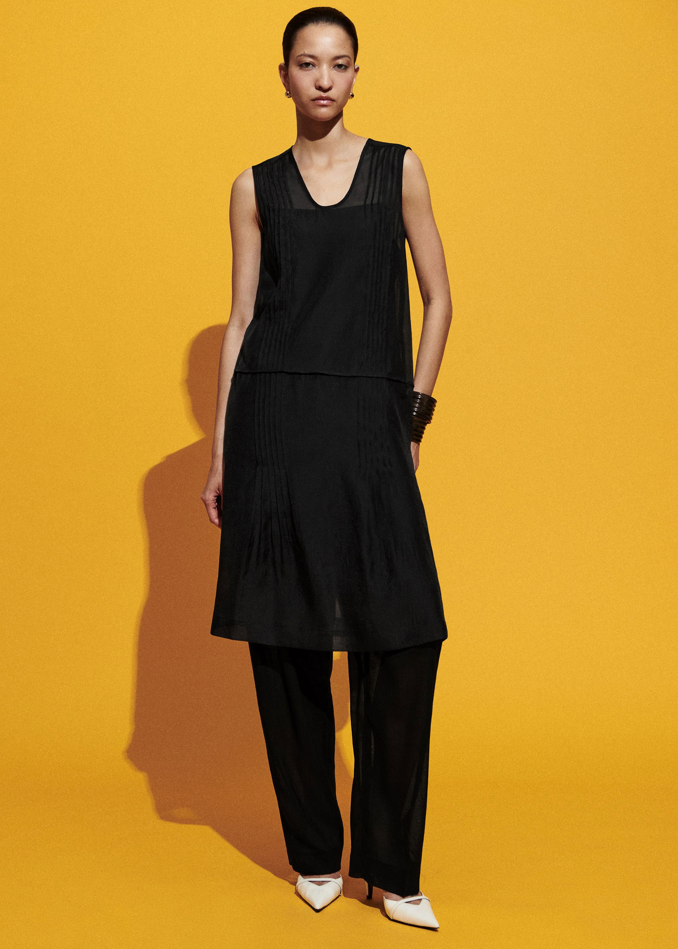 A person models a sleeveless black dress with matching pants and white heels against a vibrant yellow background.