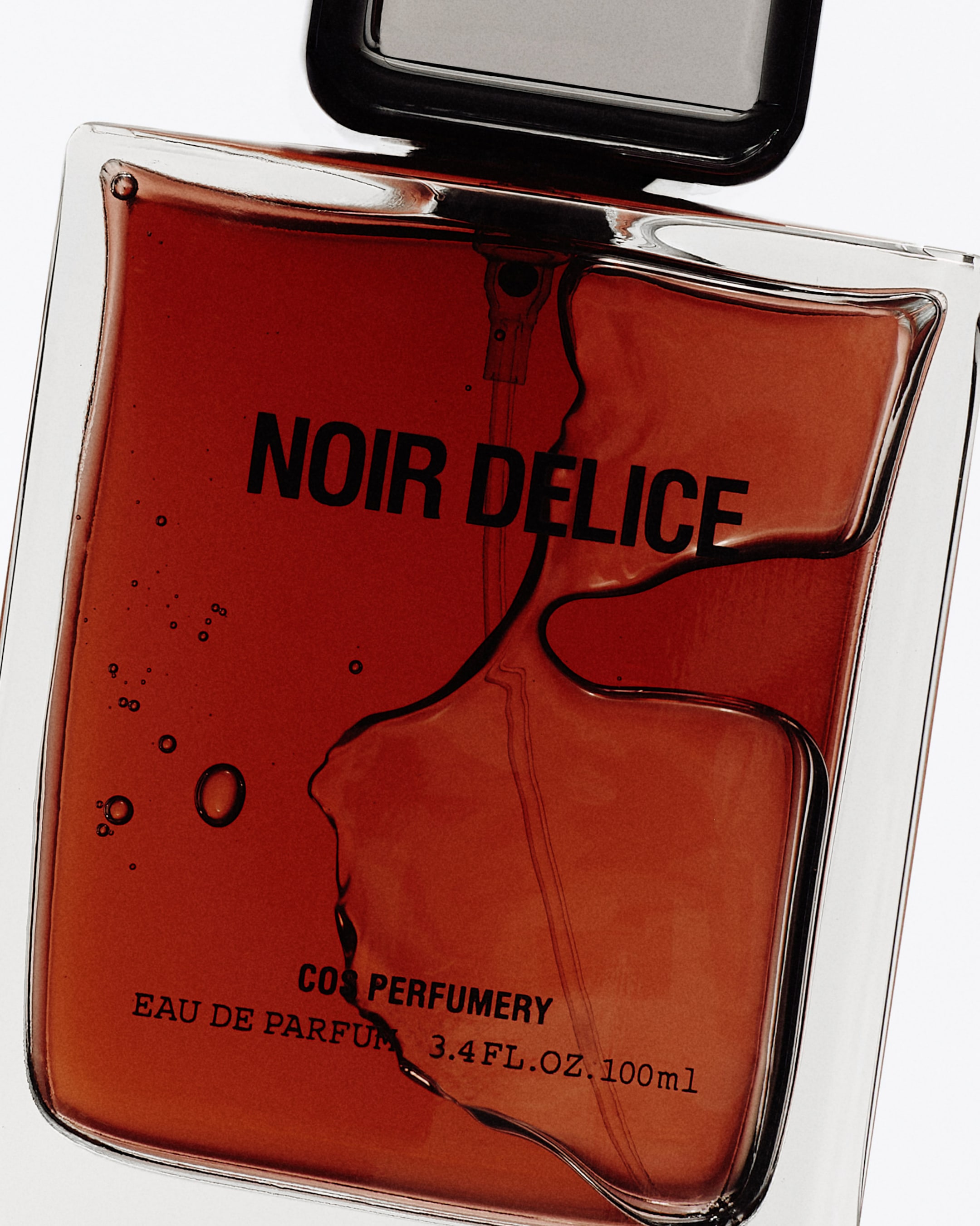 A rectangular glass bottle of Noir Délice Eau de Parfum, filled with amber liquid, featuring a bold black cap and minimalist design.