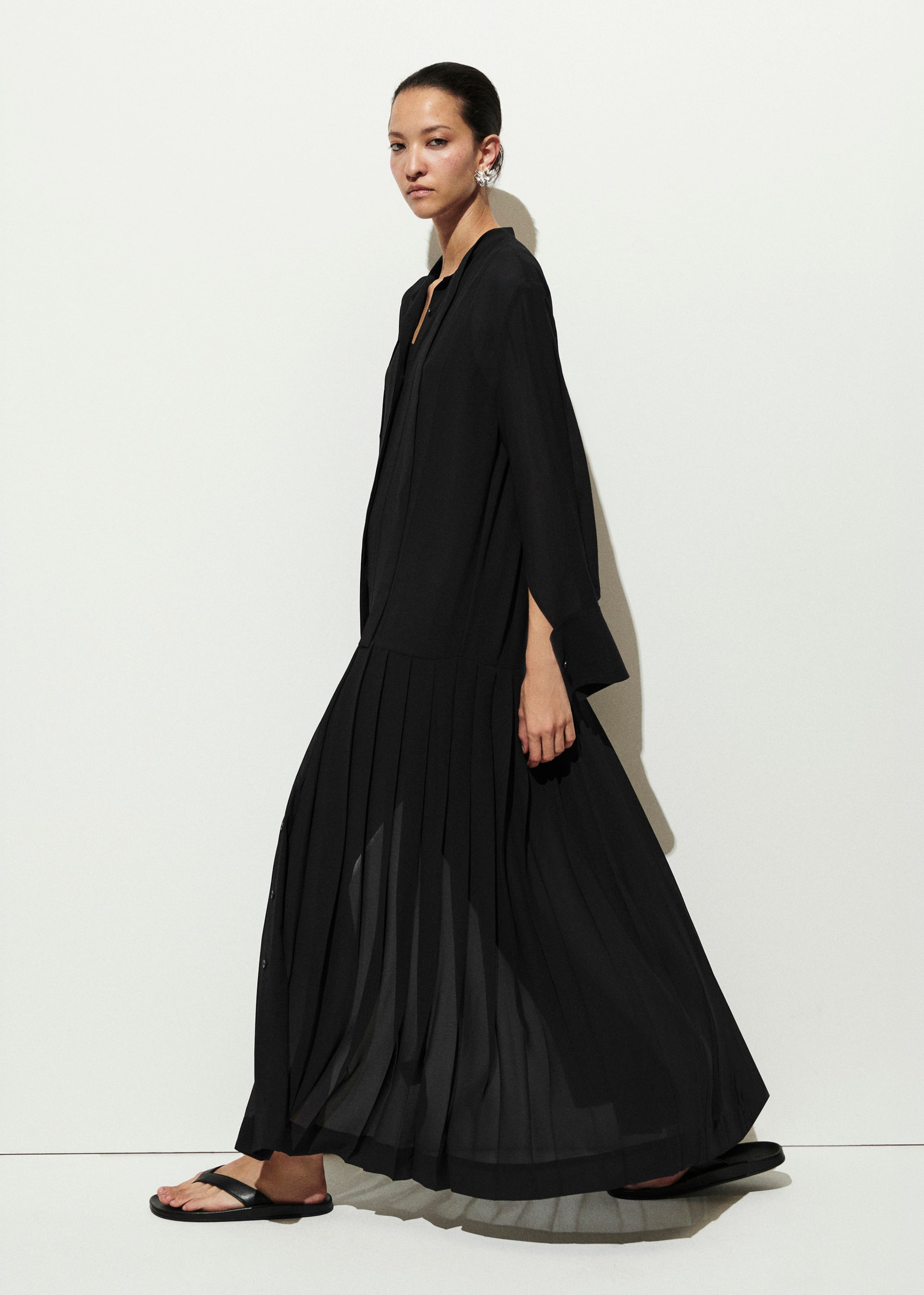 A woman in a flowing black dress and sandals poses against a plain white background, showcasing her stylish outfit and elegant posture.